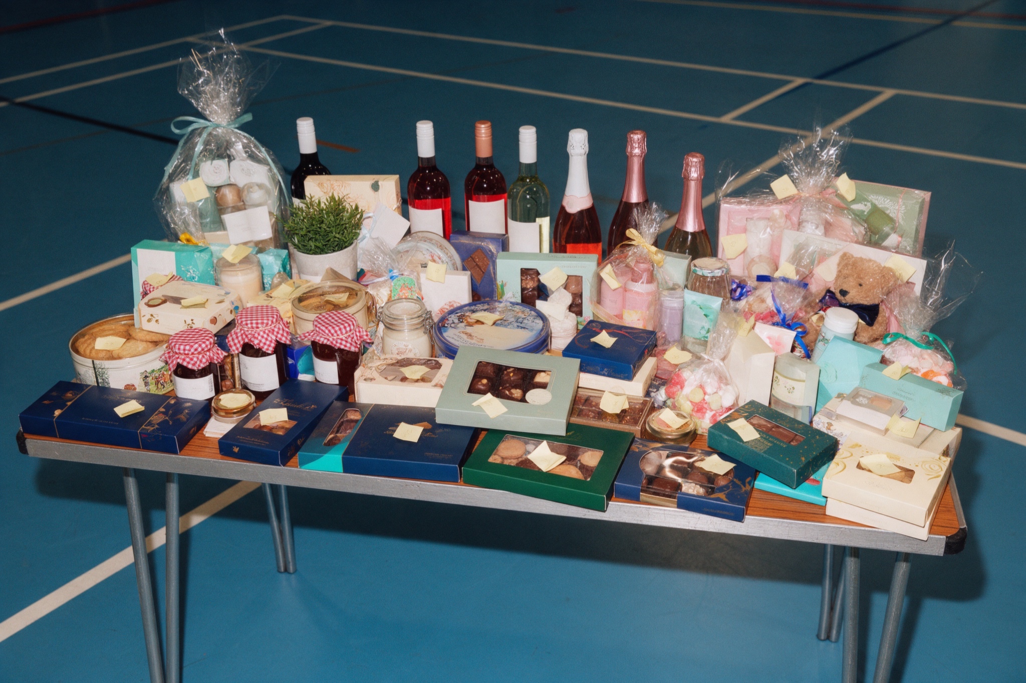 A school fair tombola table in a sports hall, crammed with chocolate boxes, jars of jam, bottles of wine, gift sets and a teddy bear, each with a small ticket attached.